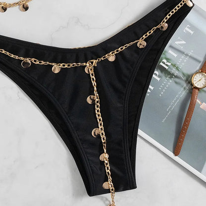 This image features the bottom of the Elise Black Bandeau Bikini. The bandeau style bikini with intricate gold chain and gold trinket accents is styled with a watch and magazine.